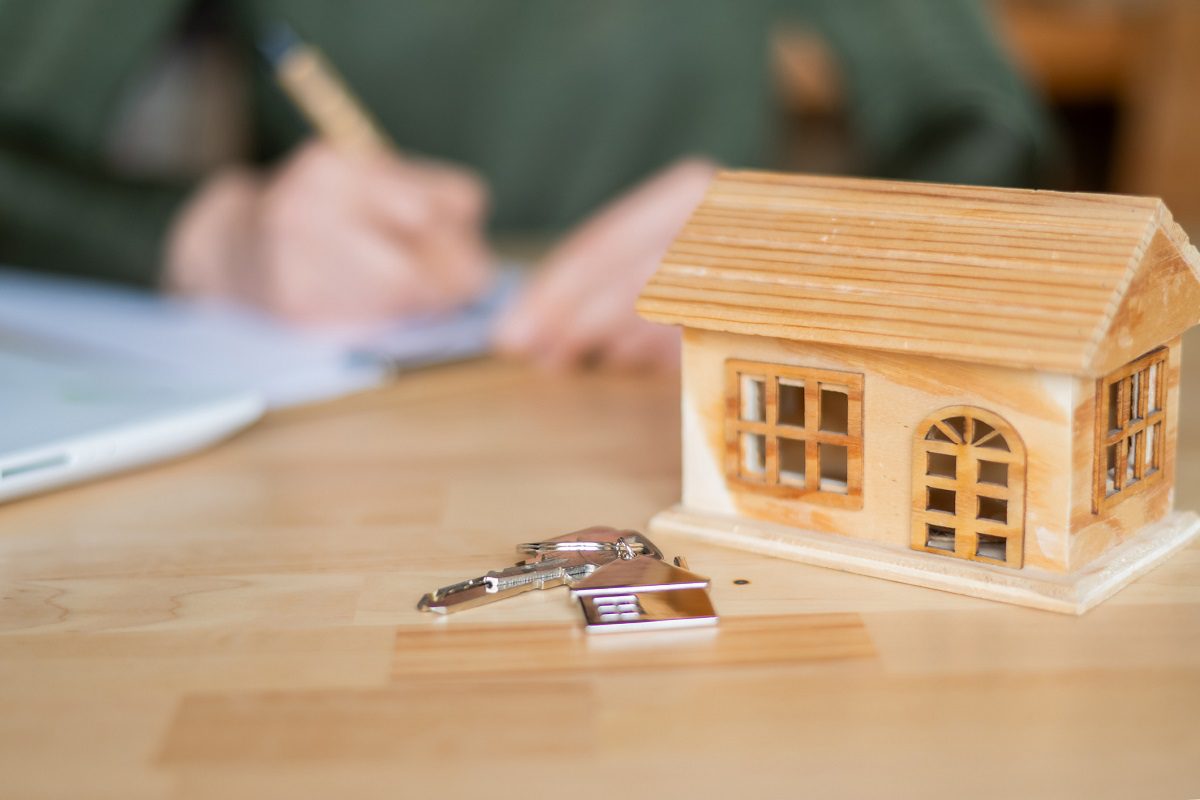 A small wooden house model and a set of keys on a table, with a person writing in the background.