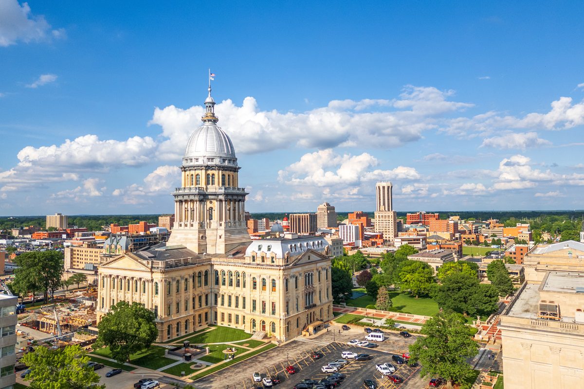A large domed government building stands in the foreground, surrounded by city buildings, trees, and a partly cloudy sky.
