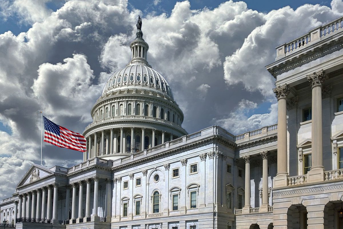 United States Capitol building with the dome and an American flag visible, set against a dramatic cloudy sky.