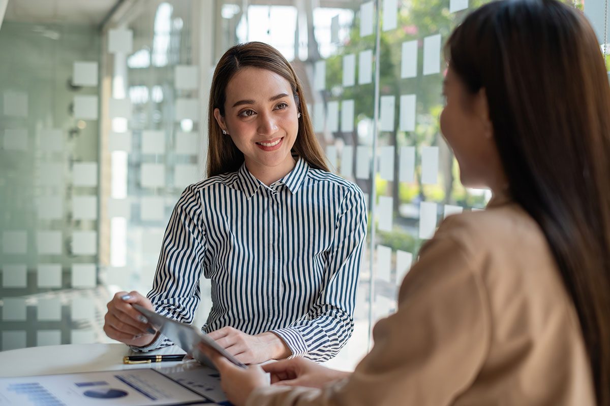 Two women sit across from each other at a table in an office, discussing documents. One woman is smiling and holding papers, while the other is partially visible from behind.