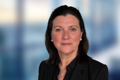 Woman with dark hair wearing a black blazer and dark blouse, posing in front of a blurred blue and white background.