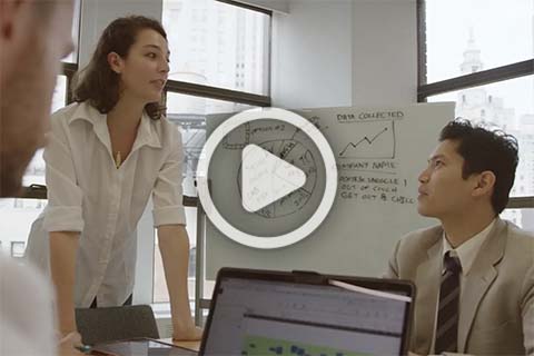 A woman stands and speaks to a seated man during a meeting in an office, with a whiteboard displaying charts and notes in the background.