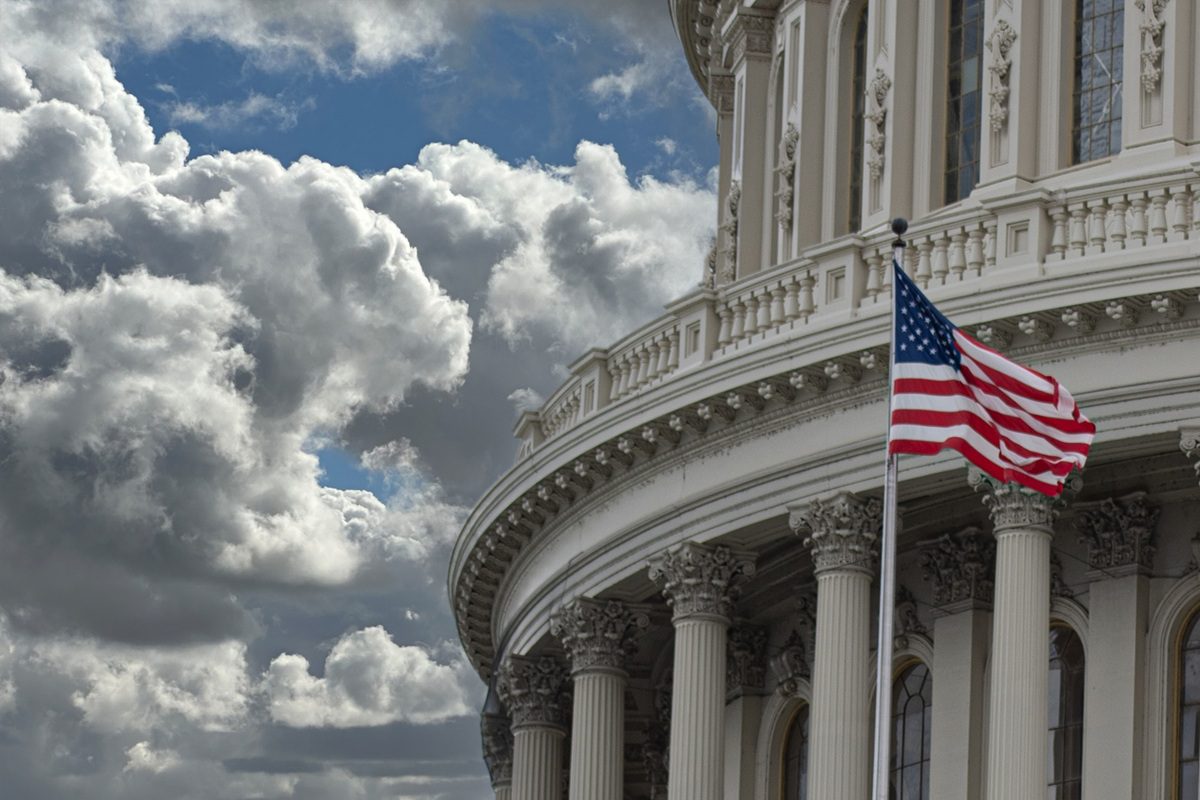 An American flag waves in front of the U.S. Capitol building under a sky with large, dramatic clouds.