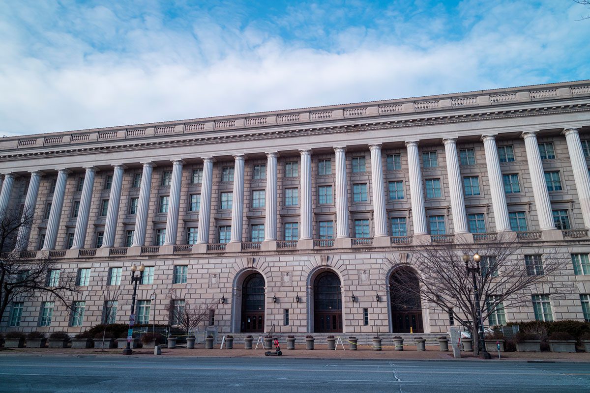 A large, neoclassical government building with tall columns, multiple windows, and an arched main entrance under a blue sky.