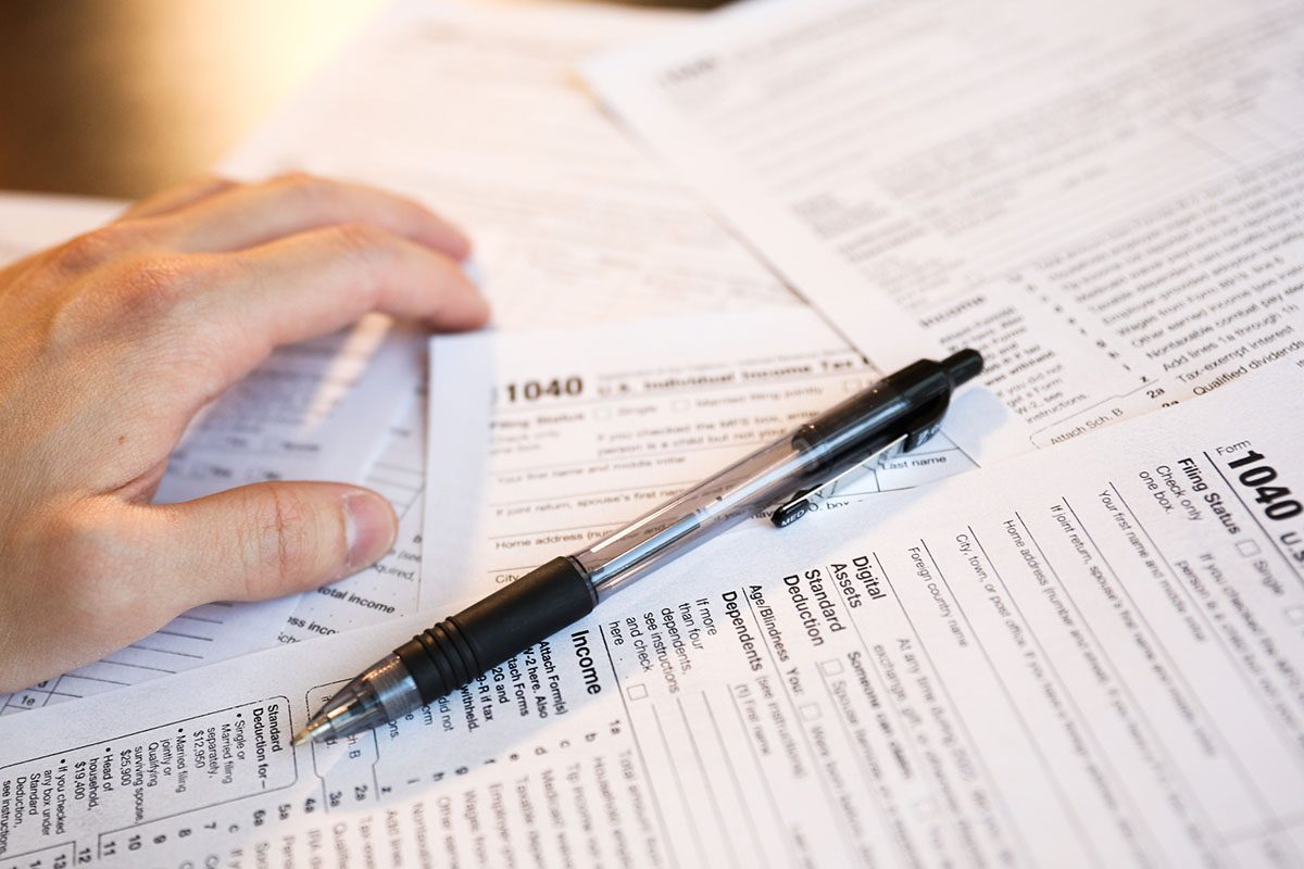 A hand rests on a stack of IRS 1040 income tax forms with a black pen placed on top of the documents.