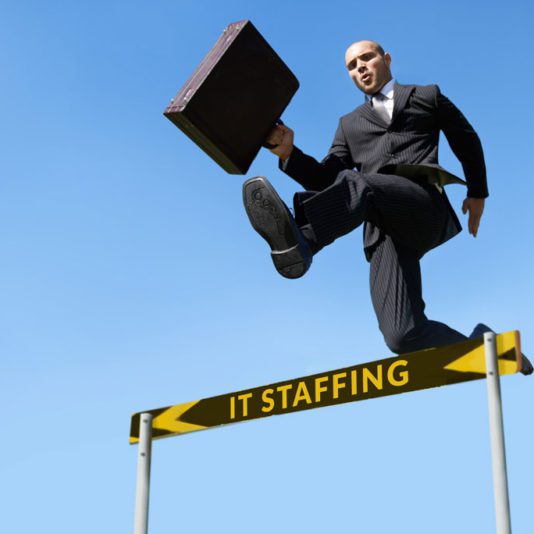 A man in a business suit jumps over a hurdle labeled IT Staffing while holding a briefcase against a clear blue sky.