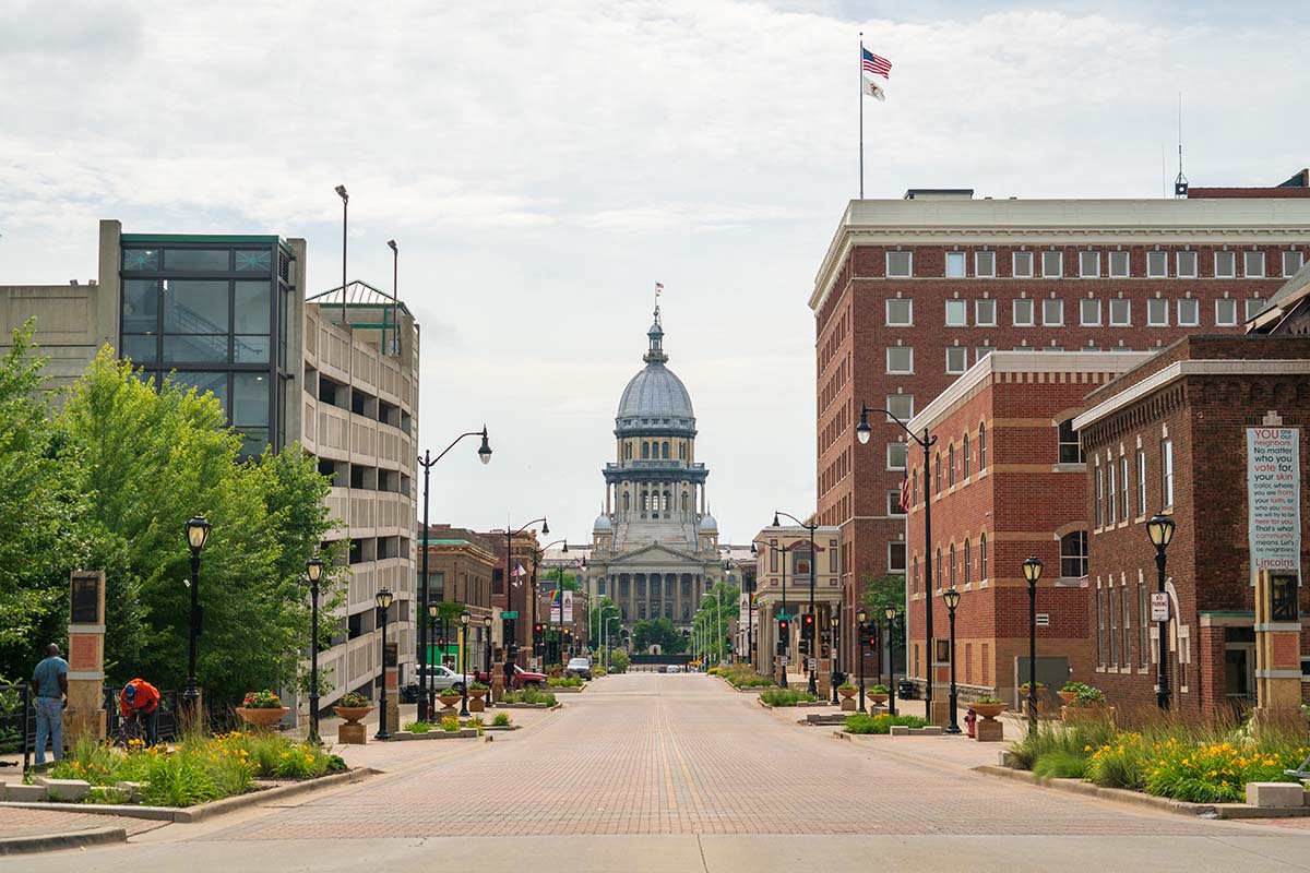 A straight street view leading to the Illinois State Capitol building, with surrounding brick and modern buildings under a partly cloudy sky.