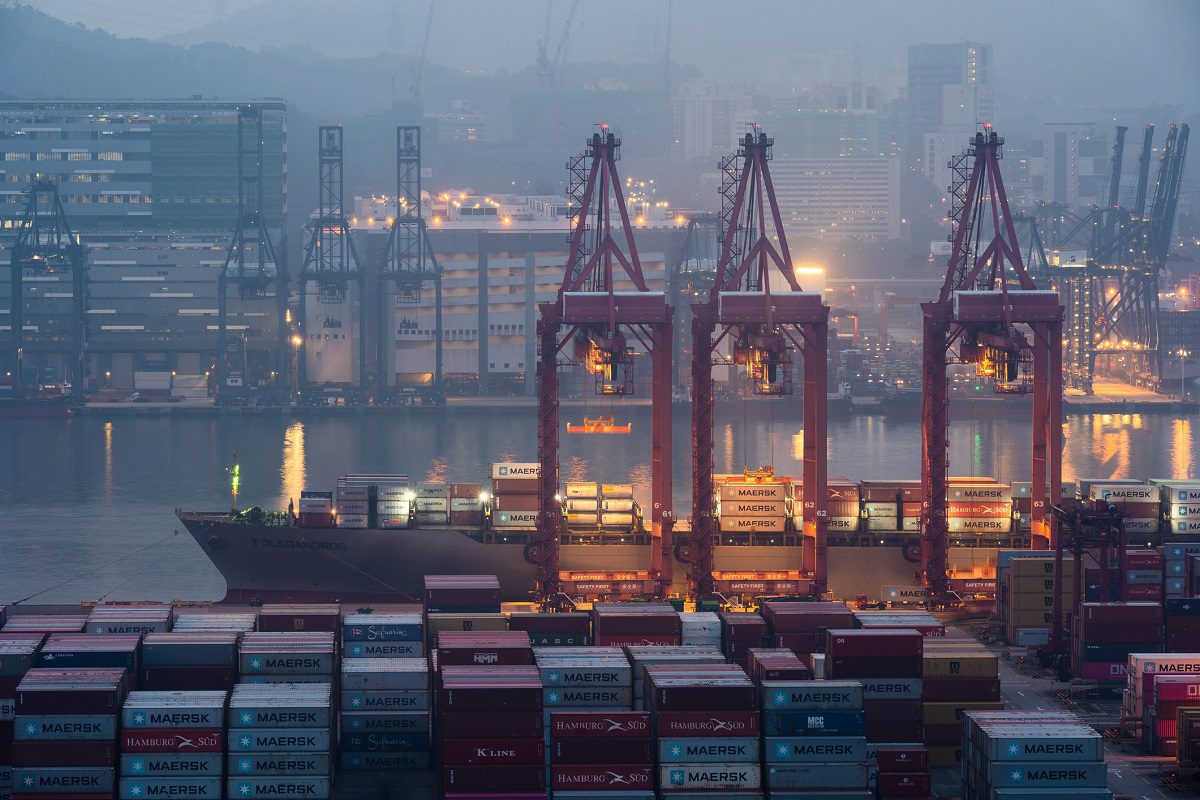Large shipping cranes load and unload containers from a cargo ship at a busy port, with stacked shipping containers and industrial buildings in the background.