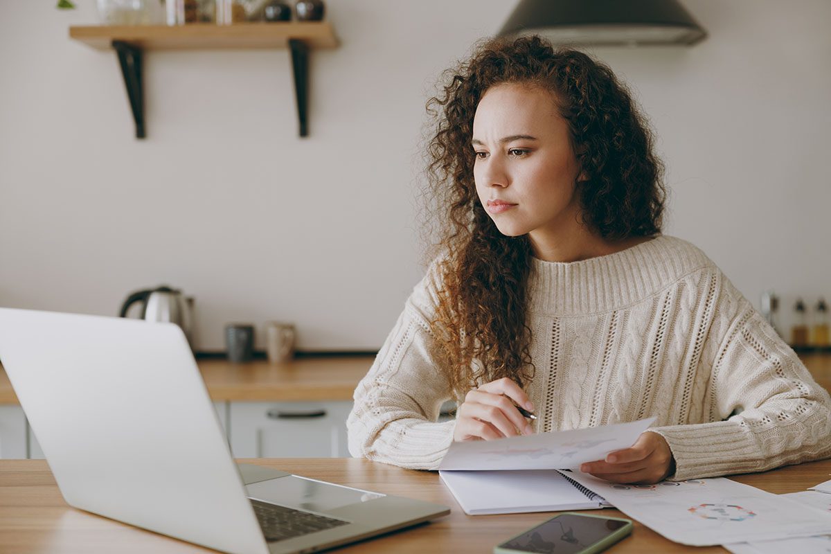 Young woman with curly hair sits at a kitchen table, looking at a laptop screen while holding papers, with a phone and documents on the table.