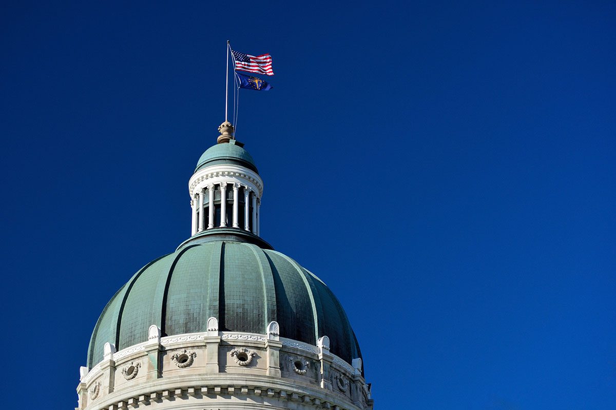 The dome of a government building with two flags, including the American flag, flying on top against a clear blue sky.
