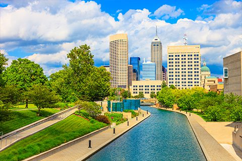 A canal runs through a landscaped park with trees, leading toward tall office buildings in a downtown city skyline under a partly cloudy sky.