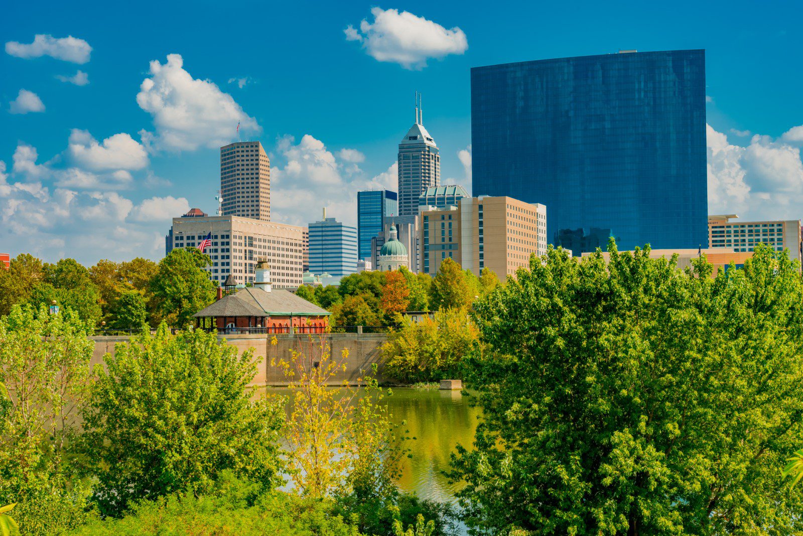 Indianapolis skyline with modern and historic buildings, trees, and a body of water in the foreground under a partly cloudy sky.