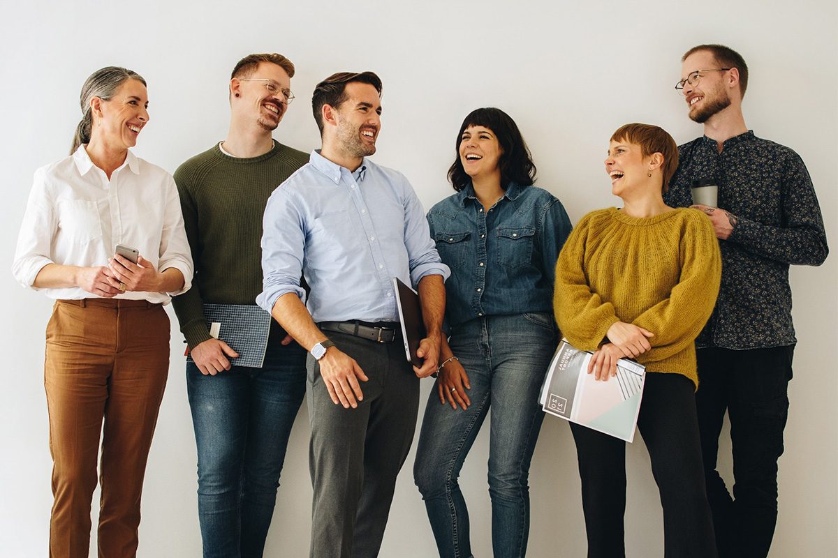 Six adults stand against a white wall, smiling and talking. Some are holding notebooks or folders, and one has a mug. They appear to be in a casual work or meeting environment.