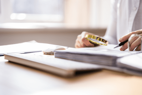 Person holding a magnifying glass and pen, examining documents on a desk with an open laptop nearby.