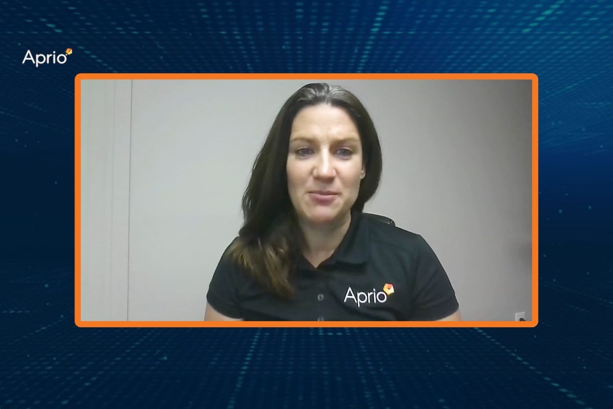 A woman wearing a black Aprio polo shirt sits in an office, framed by an orange border on a digital blue background with the Aprio logo in the top left corner.