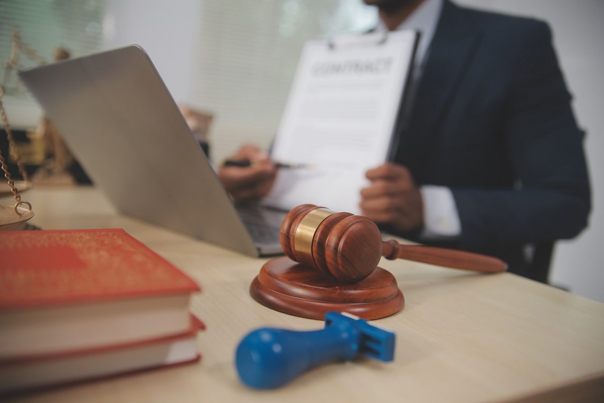 A person in a suit sits at a desk with a laptop, legal books, a gavel, a blue stamp, and holds up a clipboard displaying a contract.