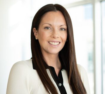 A woman with straight brown hair, wearing a white blazer over a black top, smiles while standing in a bright indoor setting.