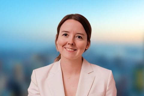 A woman with brown hair in a low ponytail, wearing a white blazer, smiles at the camera with a blurred cityscape background.