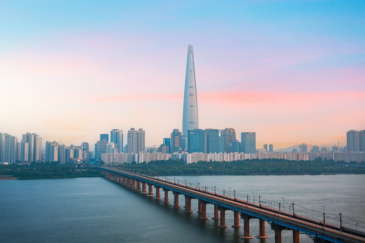 A wide river with a long bridge leads to a modern city skyline dominated by a tall skyscraper under a pastel-colored sky.