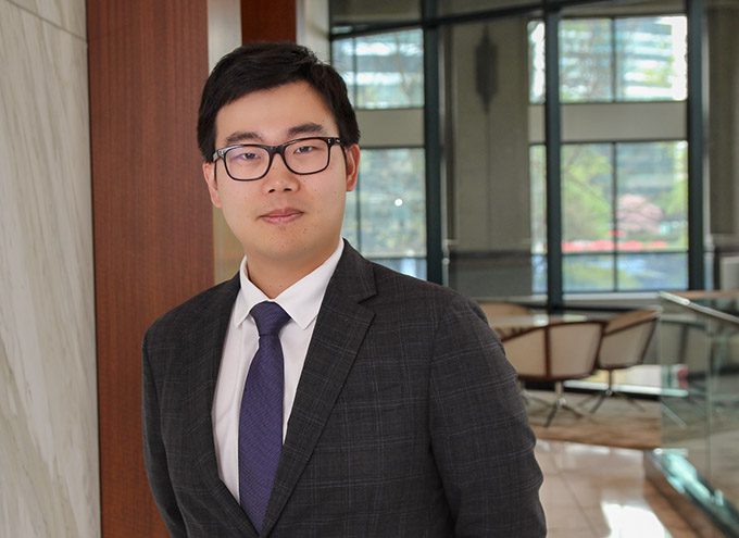 A man in a dark suit, white shirt, and purple tie stands indoors near large windows and modern office furniture.