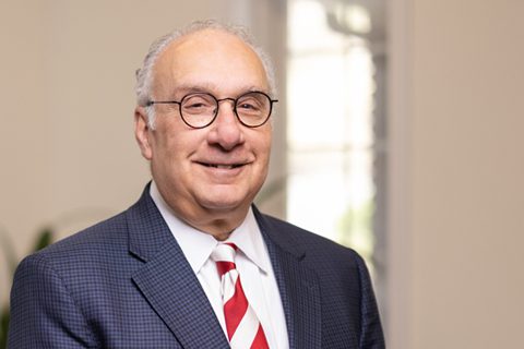 An older man with glasses and a gray suit jacket, white shirt, and red-striped tie stands in a well-lit room, smiling at the camera.