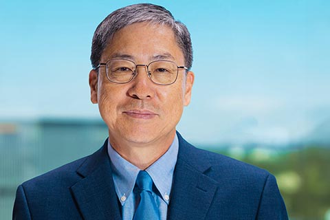 A middle-aged man with short gray hair and glasses, wearing a blue suit, light blue shirt, and blue tie, poses in front of a blurred outdoor background.