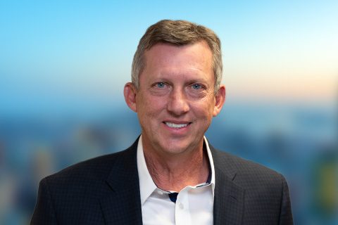 Man in a black blazer smiles in front of a bright window with a blurred city view.