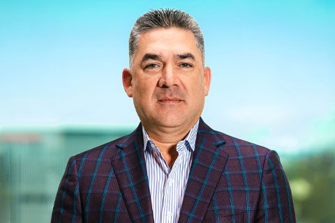 Man with short gray hair wearing a plaid suit jacket and striped shirt, standing in front of a blurred outdoor background.