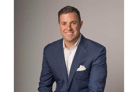 Man wearing a blue suit with a white shirt and pocket square, sitting and smiling in front of a plain gray background.