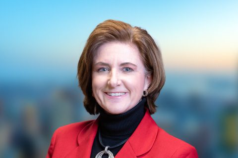 Woman with short brown hair and a red blazer smiles on a bright blurred background