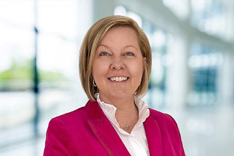 Blonde woman in a pink blazer smiles in front of a bright blurred lobby background