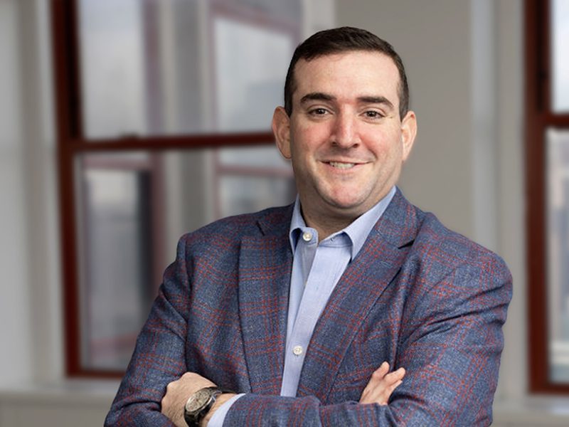 A man in a blue plaid blazer and light blue shirt stands indoors with folded arms, smiling at the camera. Large windows are visible in the background.