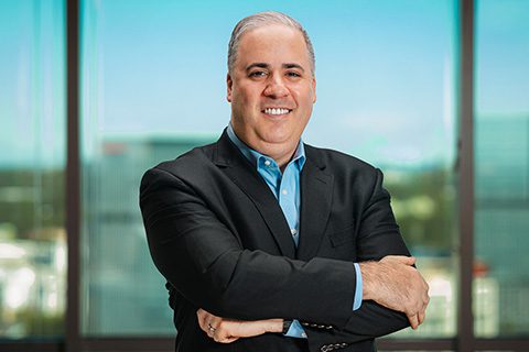 Man in a black blazer smiles in front of a bright window with a blurred city view.