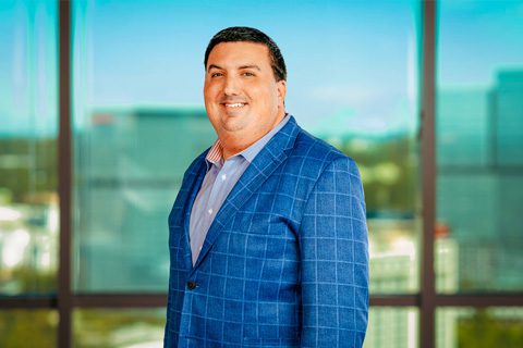 A man in a blue checked suit stands indoors in front of large windows with a cityscape view in the background.