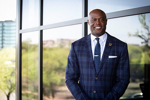 A man in a blue checked suit stands smiling in front of large windows with trees and buildings visible outside.