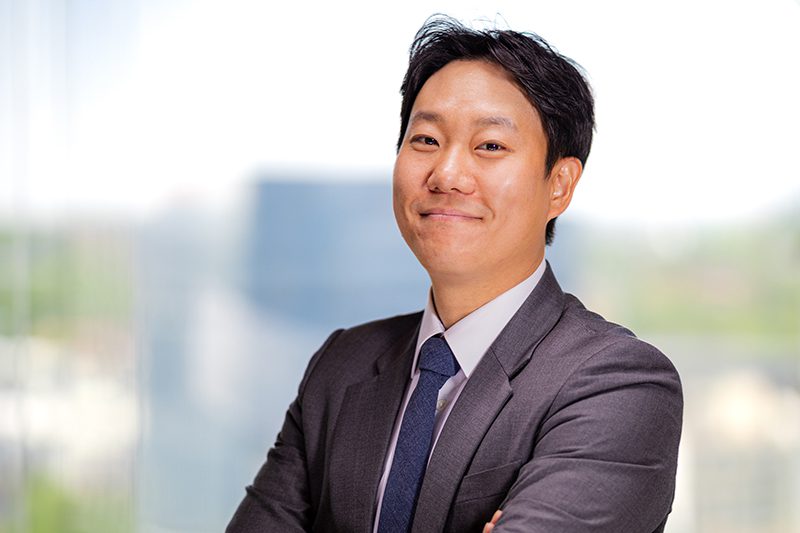 A man in a suit and tie stands with arms crossed, smiling, in front of a blurred background of buildings and greenery.