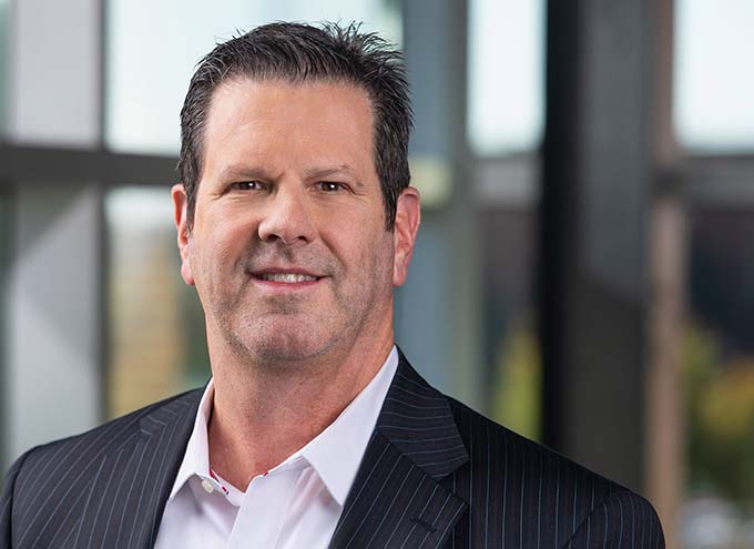 A middle-aged man wearing a dark pinstripe suit and white shirt stands indoors in front of large windows, looking at the camera and smiling slightly.