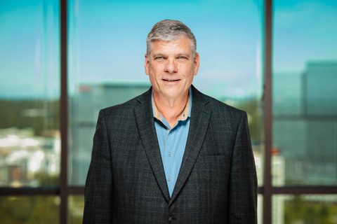 A middle-aged man wearing a dark blazer and blue shirt stands indoors in front of large windows with a cityscape background.