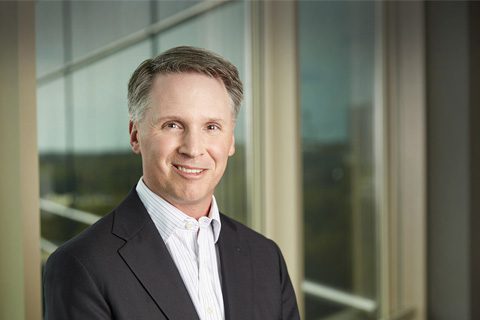 A man in a suit jacket and collared shirt stands indoors near large windows, smiling at the camera.
