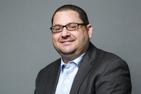 Man wearing glasses, a light blue shirt, and a dark gray suit jacket, smiling, posed in front of a plain gray background.