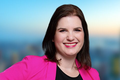 Dark haired woman in a fuschia blazer smiles in front of a bright window with a blurred city view