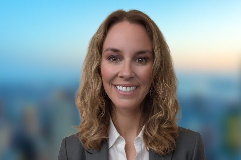 A woman with wavy light brown hair, wearing a gray blazer and white shirt, smiles in front of a blurred cityscape background.