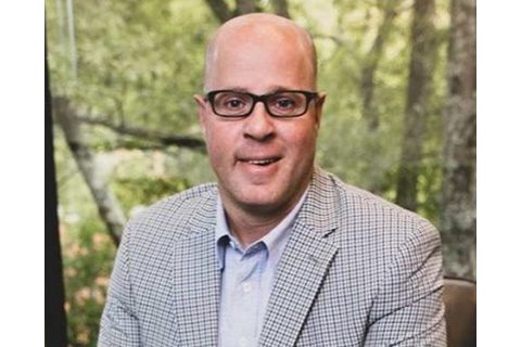 A man with glasses and a checked blazer sits indoors in front of a window with a green, leafy outdoor background.
