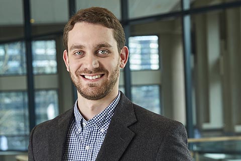 A man with short brown hair and a beard, wearing a checked shirt and dark blazer, stands indoors in front of large windows.