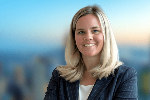 A woman with blonde hair in a pinstripe suit jacket smiles with arms crossed, standing in front of a blurred cityscape background.