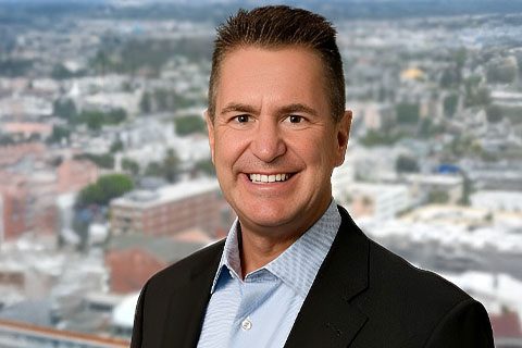 Dark haired man in a blazer smiles in front of a blurred city background
