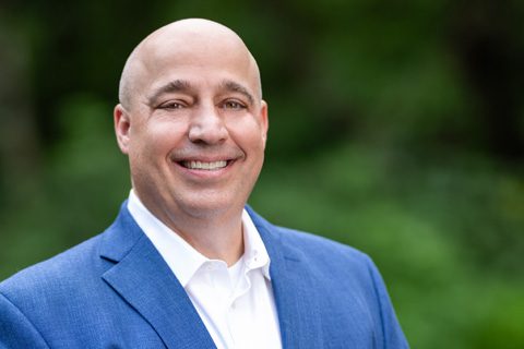 Bald man in a blue suit jacket and white shirt smiling outdoors in front of a blurred green background.