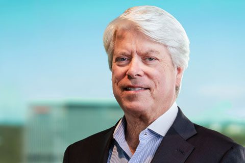 A middle-aged man with white hair in a dark suit jacket and light blue shirt stands in front of a blurred office building background.