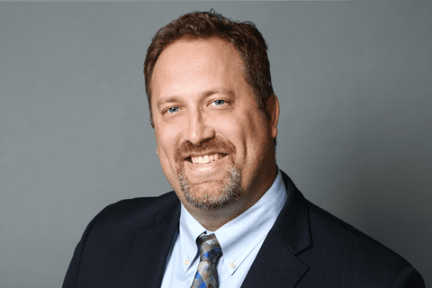 A man with short brown hair, a beard, and a mustache wearing a dark suit, light blue shirt, and patterned tie poses in front of a plain gray background, smiling at the camera.