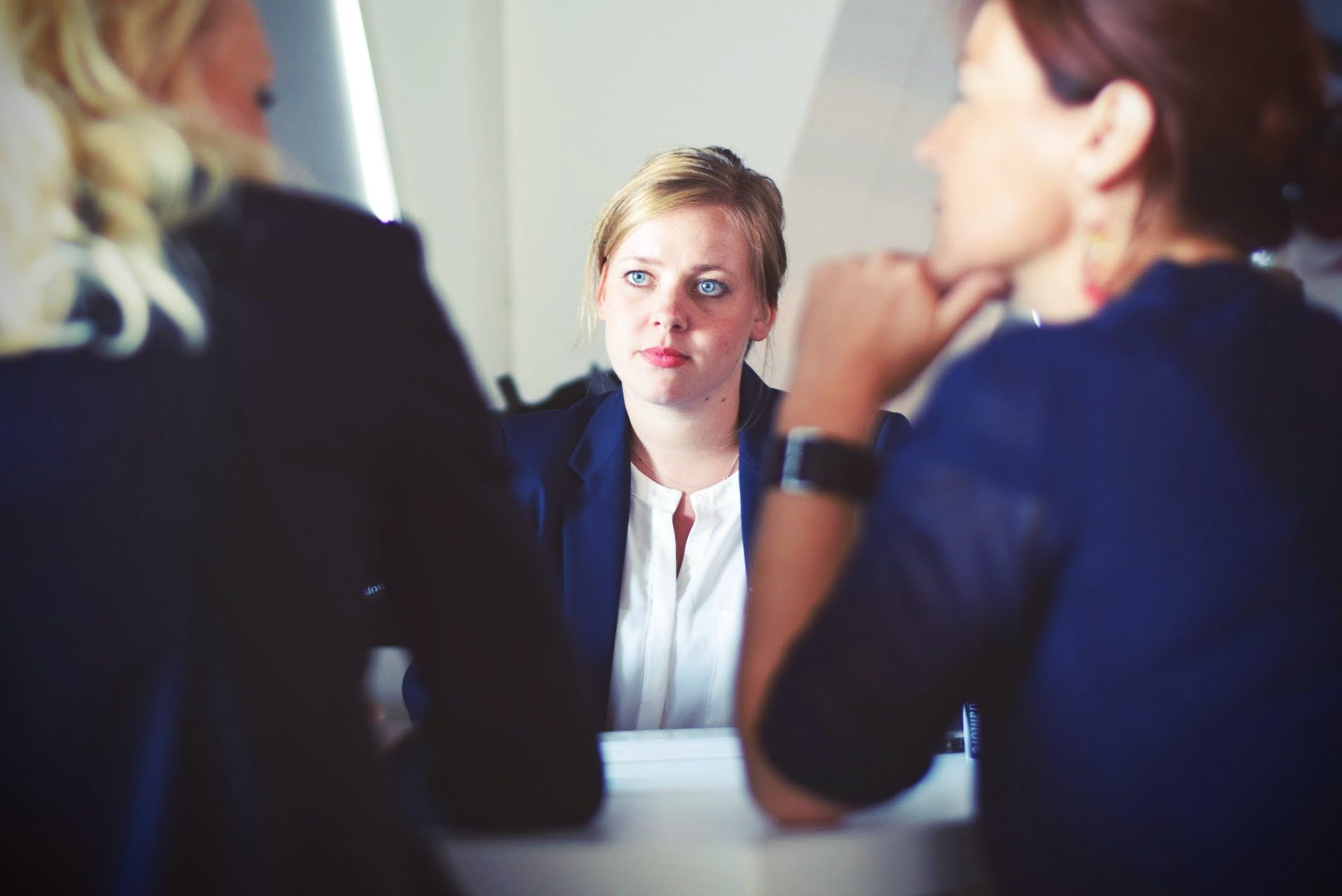 Three women in business attire sit at a table, with one woman facing the camera and two others seen from behind, suggesting a professional meeting or interview setting.